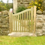 Handcrafted wooden gate by Richard Hope Wooden Gates, featuring natural timber with a curved top design, installed between stone pillars for a rustic garden entrance.