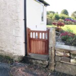 Wooden garden gate with vertical slats, attached to a stone wall beside a house exterior.