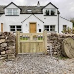 Handcrafted wooden gate at a traditional white cottage with stone walls, featuring bespoke design, quality timber, and rustic charm, perfect for enhancing curb appeal and security.