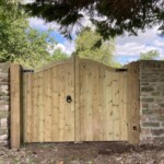 Handcrafted wooden garden gate with a natural finish, sturdy iron hinges, and decorative ring handle, set in a brick wall under a lush tree canopy. Perfect for enhancing garden security and aesthetic appeal.