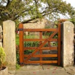 Rounded wooden garden gate with metal hinges, set between weathered stone pillars, leading to a lush garden with mature trees and stone cottages in the background. Perfect for rustic outdoor charm and privacy.